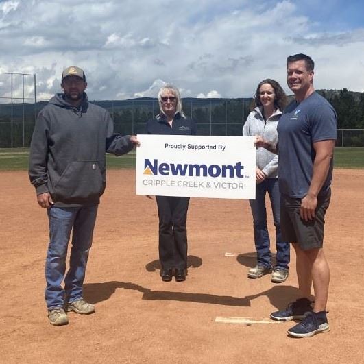 Photo of four adults in a baseball field holding a sign that says 